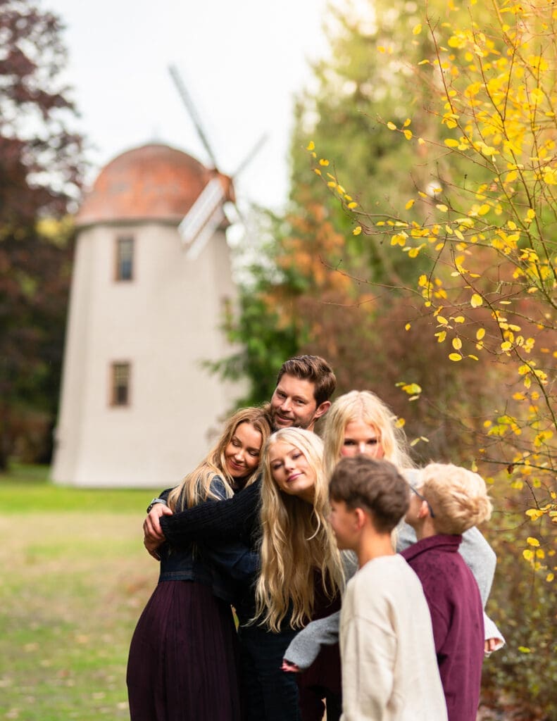 A family shares a tender moment during a fall family photoshoot at Marymoor Park in Redmond, WA. The parents embrace gently, framed by their children nearby. Behind them, the iconic Marymoor windmill rises amidst golden autumn leaves, creating a heartfelt, seasonal backdrop. This image captures authentic family connection and the timeless beauty of outdoor portraits on Seattle’s Eastside.