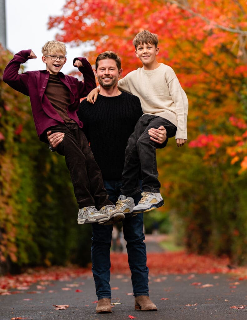 Fall family photoshoot in Redmond, WA. Father holding his sons, while one son flexes his muscles. A family showing strength and happiness.