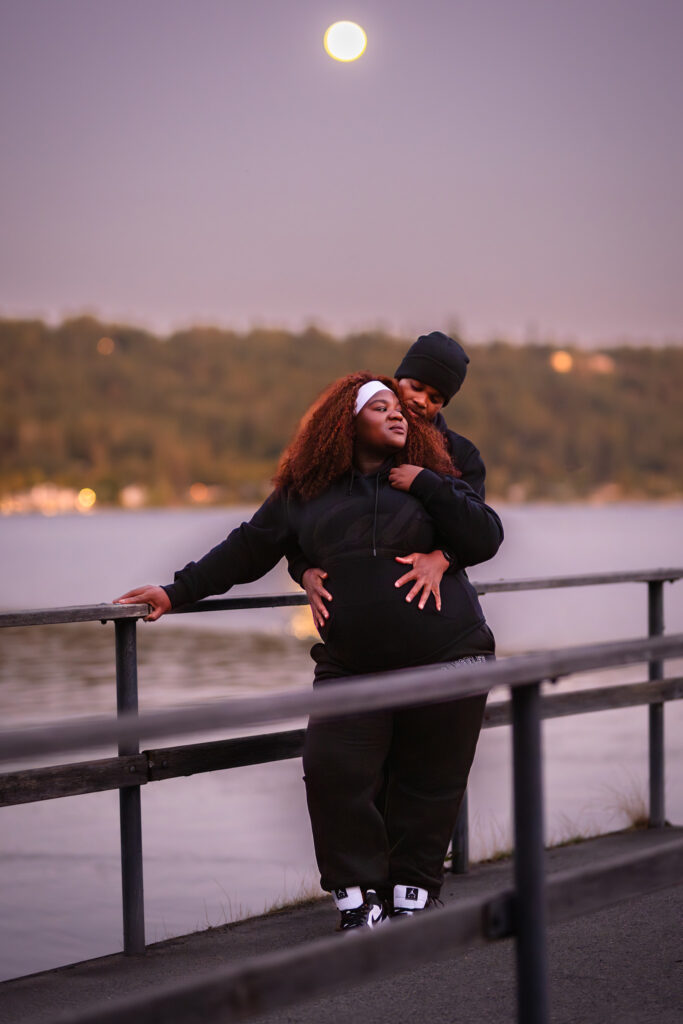 One in a series of photographs from a romantic maternity photoshoot of a couple embracing on a dock at nighttime in Redmond, WA. The full moon is reflected over the water. The couple is shown wearing black tracksuits, husband has his hands intimately wrapped around his pregnant wife's belly.