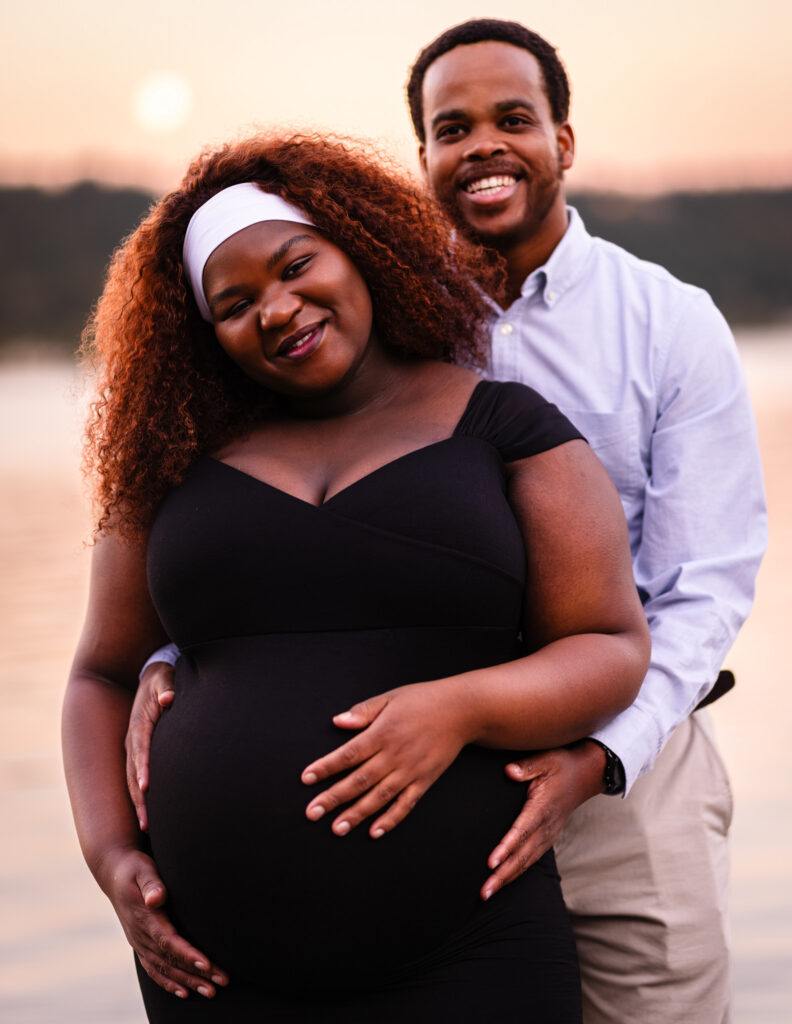 Photograph of a radiant couple during their maternity photoshoot - shown here smiling against a setting sunset whose vibrant colors are reflected off the lake water.