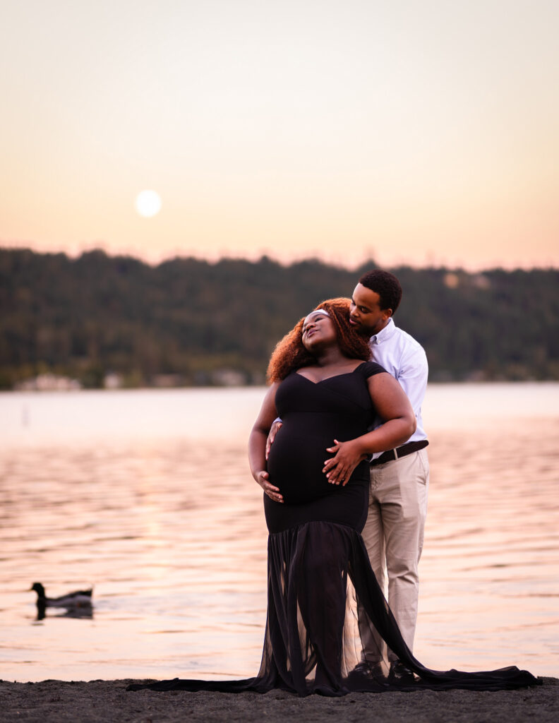 Photograph of a couple embracing during their maternity photoshoot in Seattle's Eastside. Husband is shown with his hands wrapped around his wife's pregnant belly, their hands intertwined in a romantic embrace. Behind them is a glowing, setting sun descending the golden sky - whose vibrant colors are in turn reflected off Lake Sammamish behind them. A duck has just drifted behind them and is shown about to leave the photograph frame.