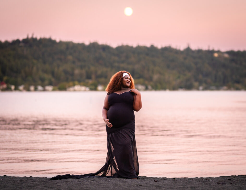 Maternity photographs of a black woman wearing an elegant black dress. The golden sun is setting behind her into the forest yet is shown glowing bright in the dark, salmon colored sky. The sunset hues are reflected off the lake water behind her.