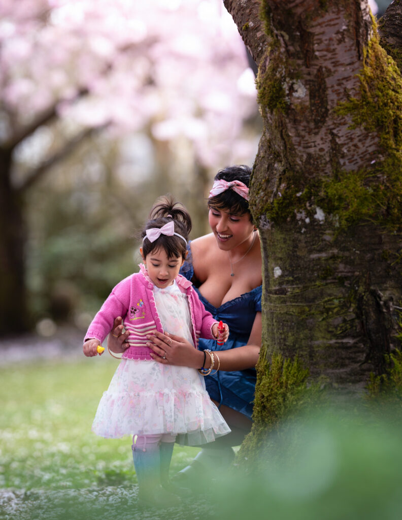 Two year old girl and her mom showing wonder at the cherry blossom petals on the ground. Behind them is a cherry blossom tree in full bloom.