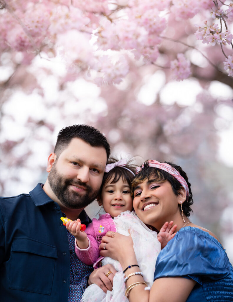 A family of three are shown smiling and hugging against a beautiful cherry-blossom backdrop