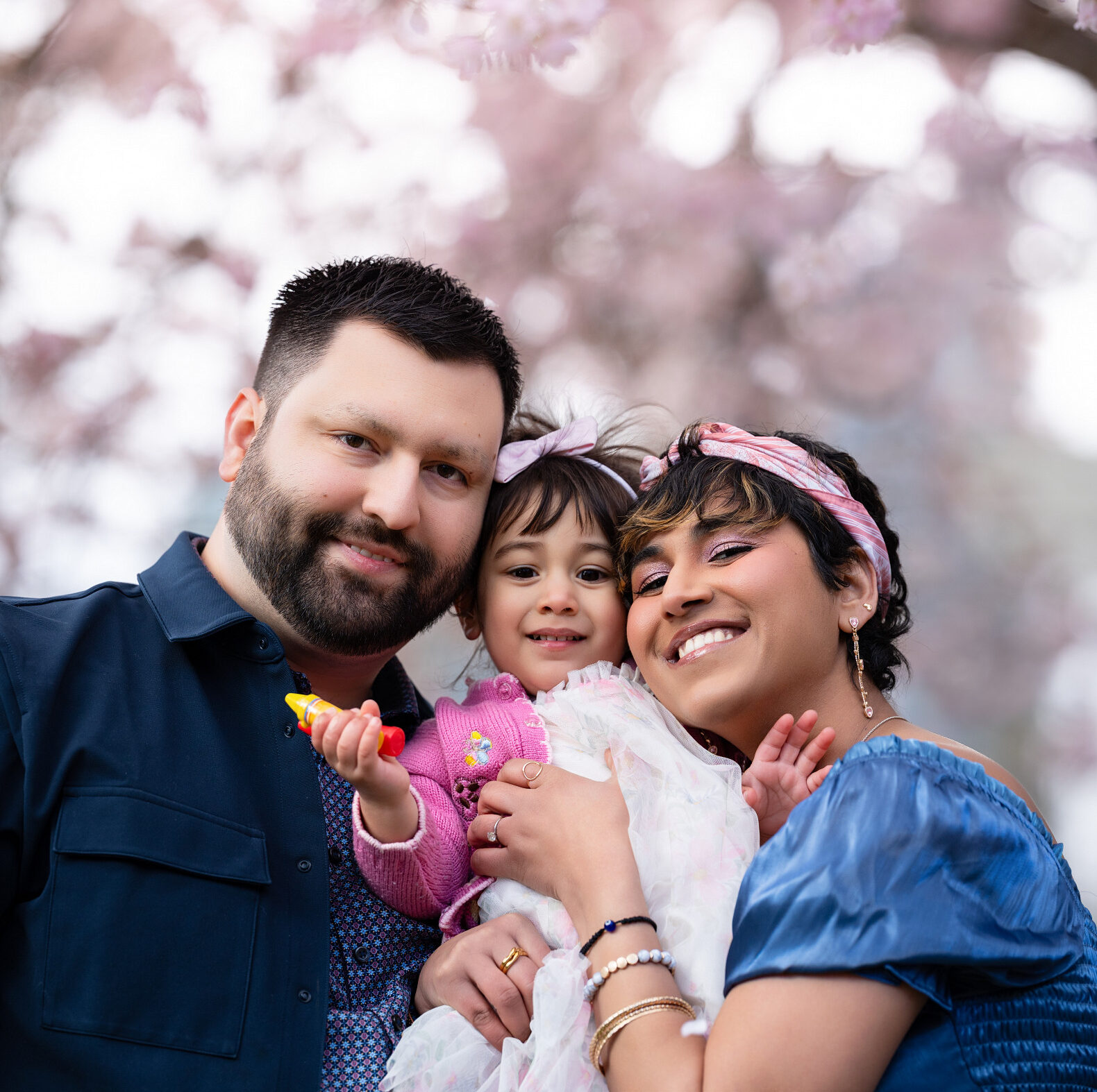 A family of three are shown smiling and hugging against a beautiful cherry-blossom backdrop
