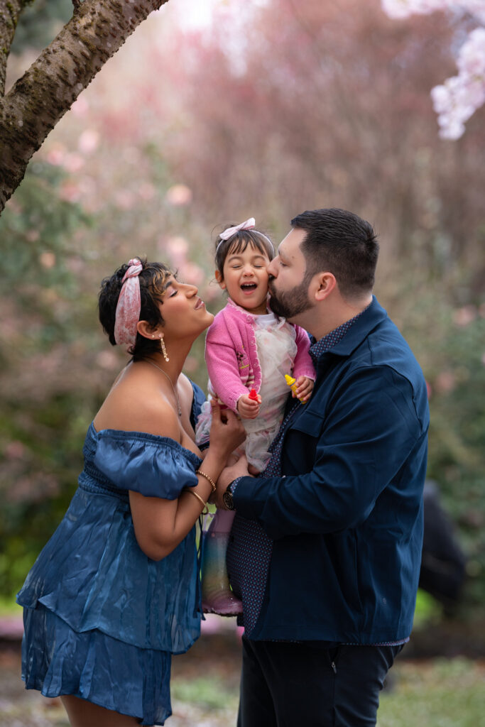 Parents kissing their daughter against a stunning backdrop of cherry blossom branches displayed with incredible bokeh, giving a dreamy quality to this photograph. Photograph was taken in Bellevue Downtown Park.