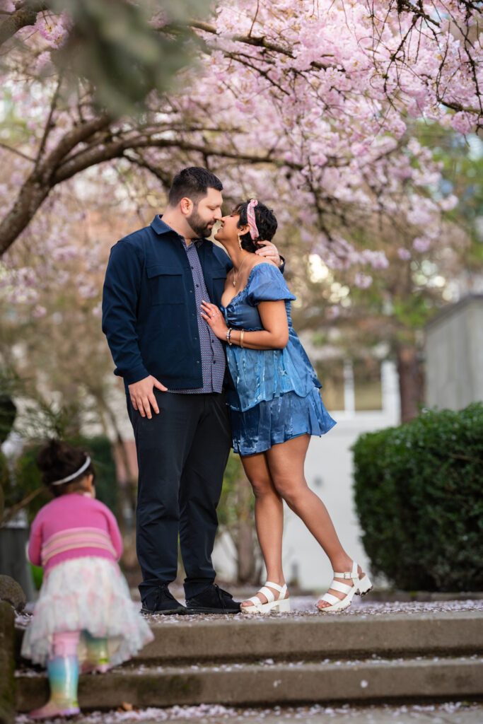 Parents embracing, nose to nose, under cherry blossom arches as their daughter explores nearby, during their family photoshoot in Bellevue WA.