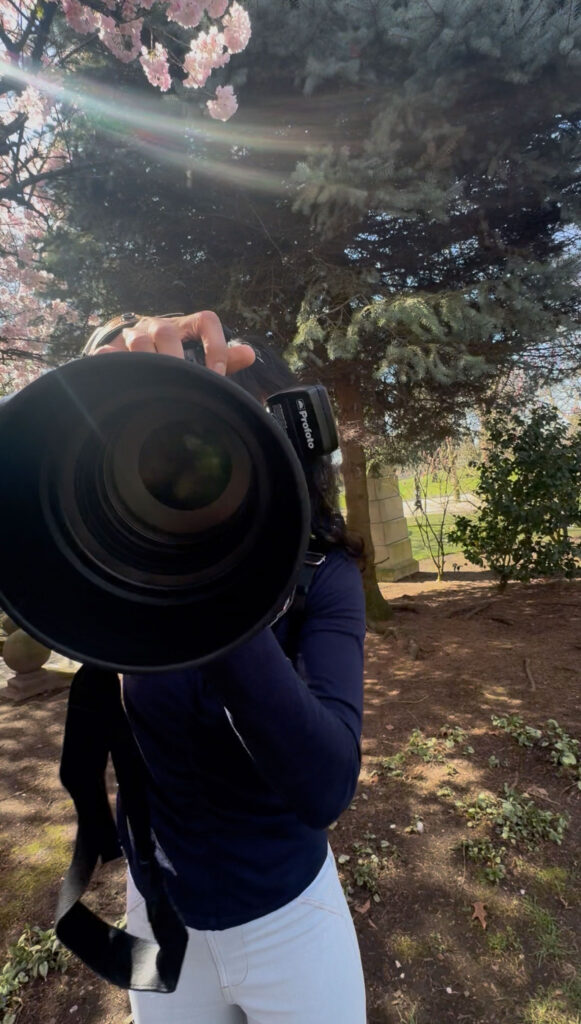 Photographer with her 135mm lens facing the viewer. Cherry blossom branches are peeking behind her along with some sun flare.